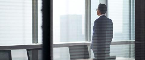 Business professional in a suit standing by office window, looking at city skyline in a modern workspace.