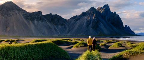 Two people walking through a grassy coastal landscape toward dramatic rocky mountains.